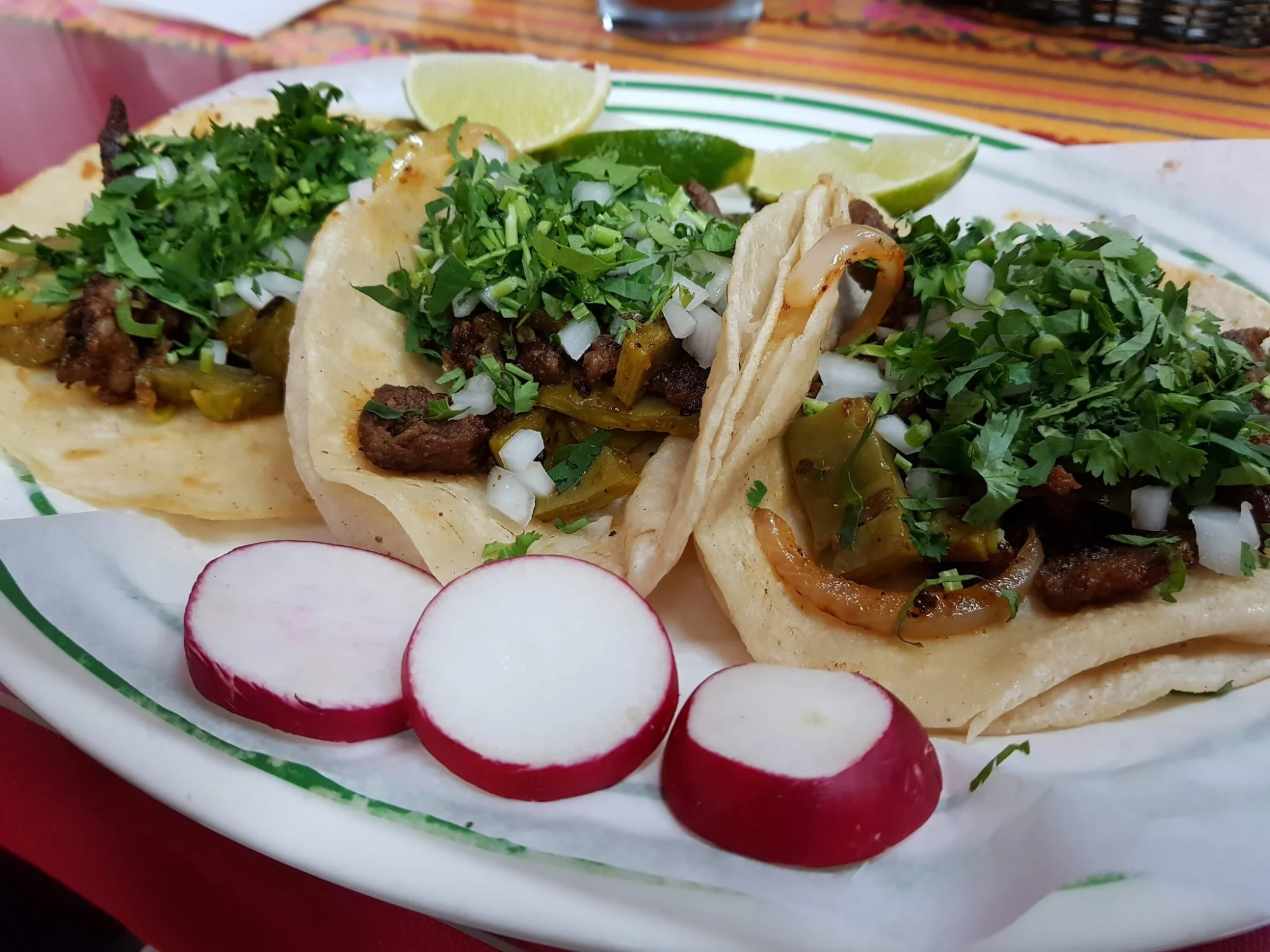 Three tacos topped with chopped greens, onions, and lime wedges on a plate at Mi Lindo San Miguelito, Kingsbridge.