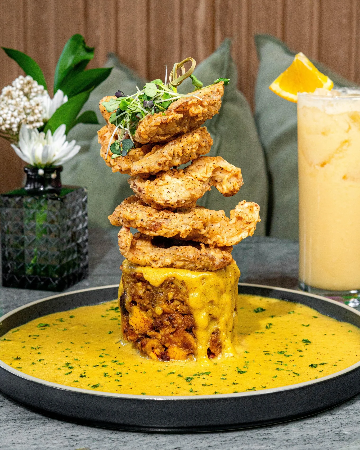 A tower of fried onions perched on a savory dish with sauce, garnished with a drink garnished with an orange slice at HAAM Caribbean Plant Cuisine, Williamsburg, Bronx NYC.
