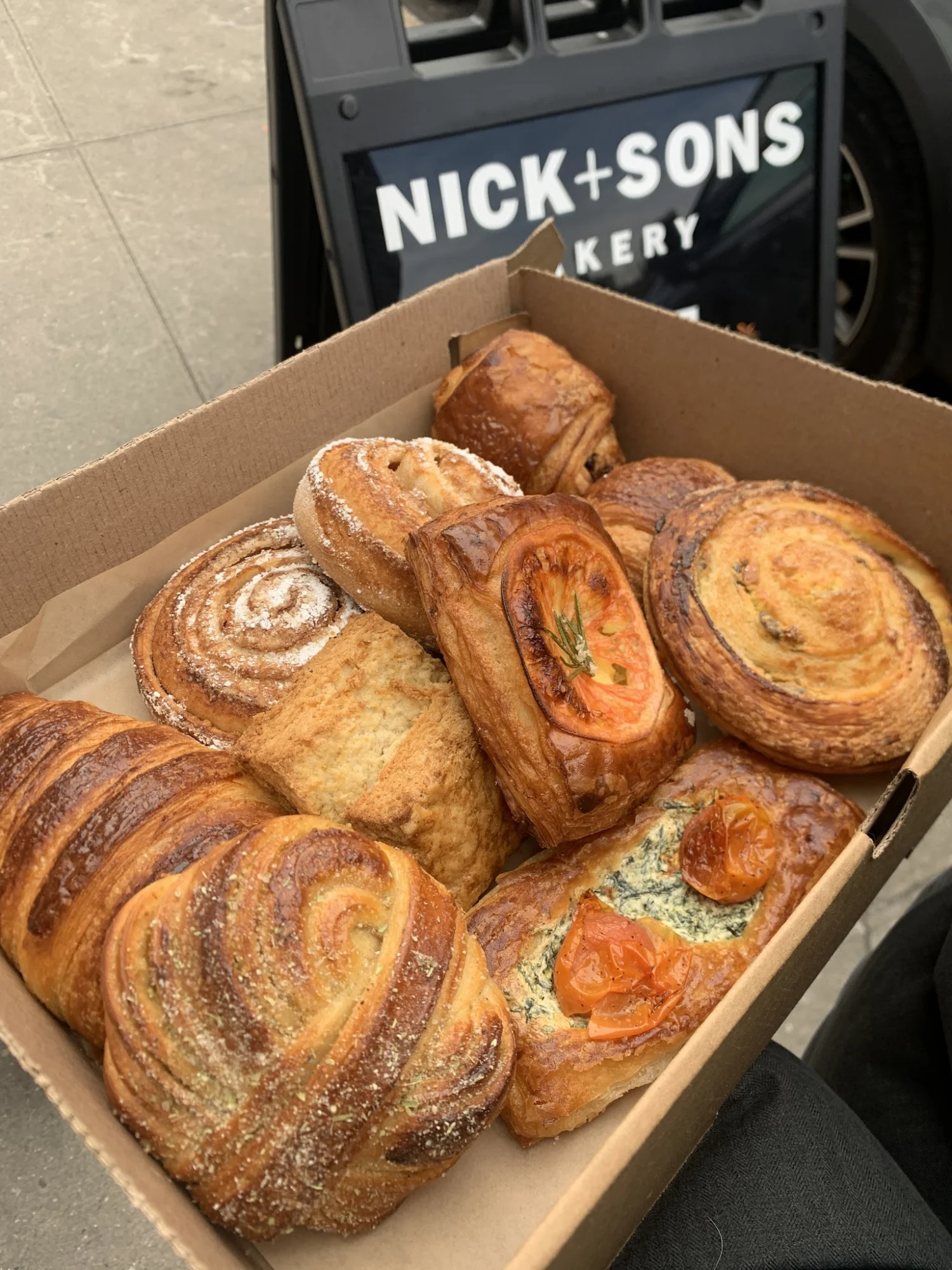 Box of assorted pastries including swirls and topped breads outside Nick + Sons Bakery, Greenpoint.