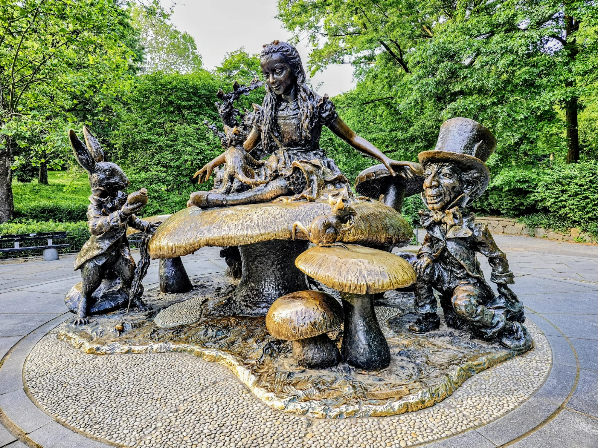 Alice in Wonderland featured image Bronze statue of Alice, Mad Hatter, and rabbit with surrounding mushrooms in a park setting, Upper East Side.