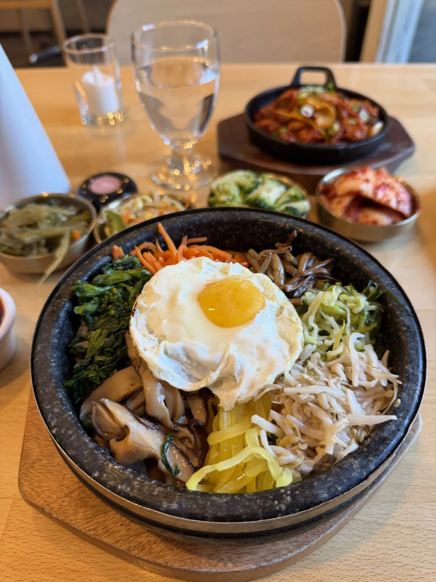 A stone bowl filled with bibimbap, topped with a fried egg and surrounded by side dishes at Kuku Korean Cuisine, Long Island City.