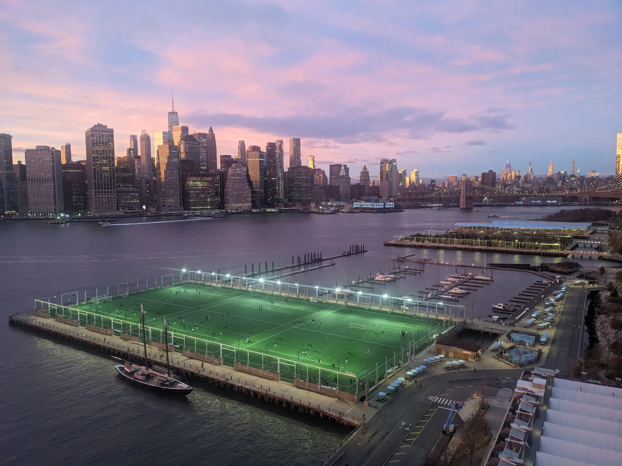 Brooklyn Bridge Park Pier 5 featured image Aerial view of Brooklyn Bridge Park Pier 5 at sunset in Brooklyn Heights, showing lit sports fields and distant city skyline.