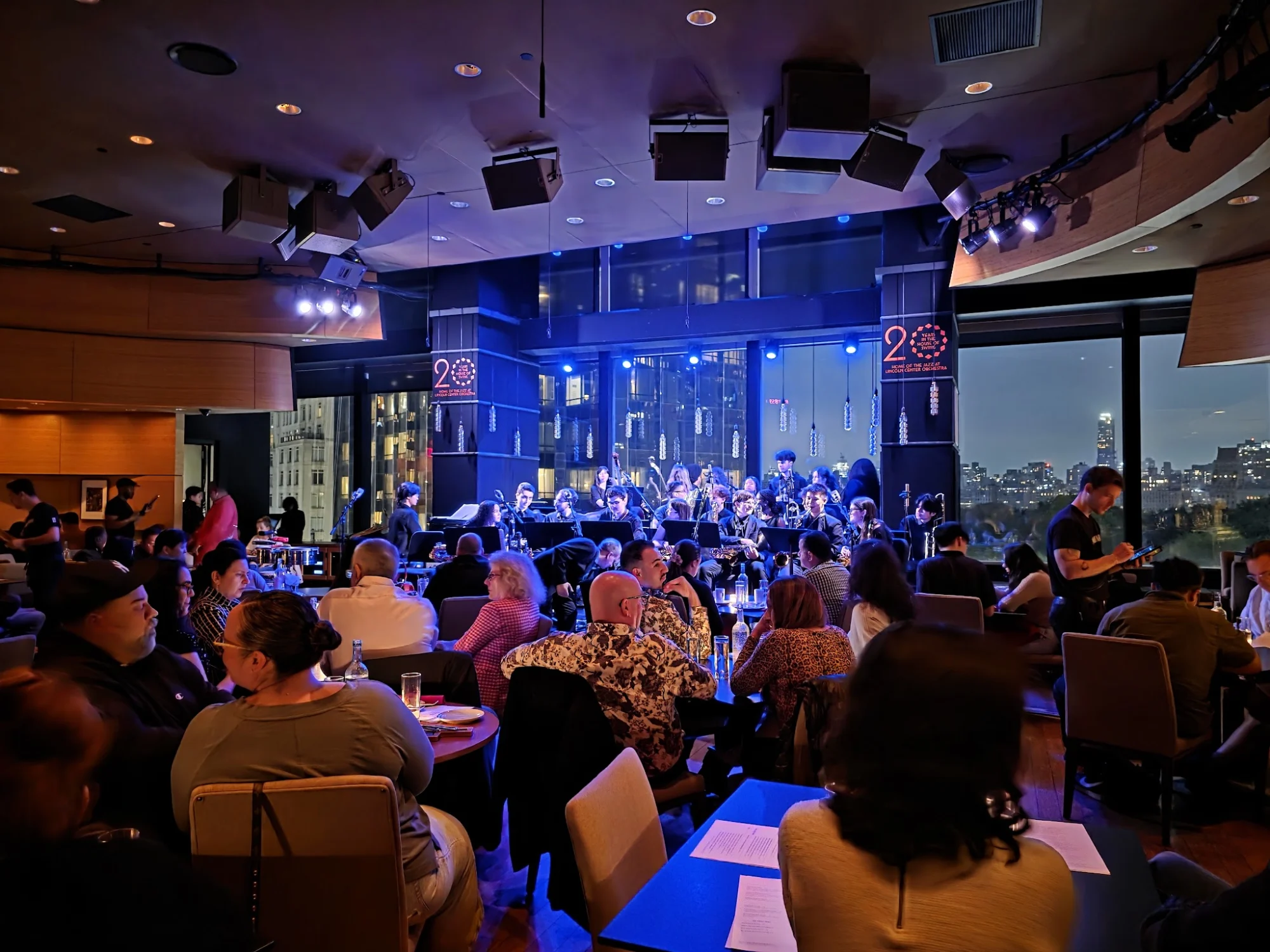 Interior of Dizzy's Club, Hell's Kitchen, with people watching a live band and city views.