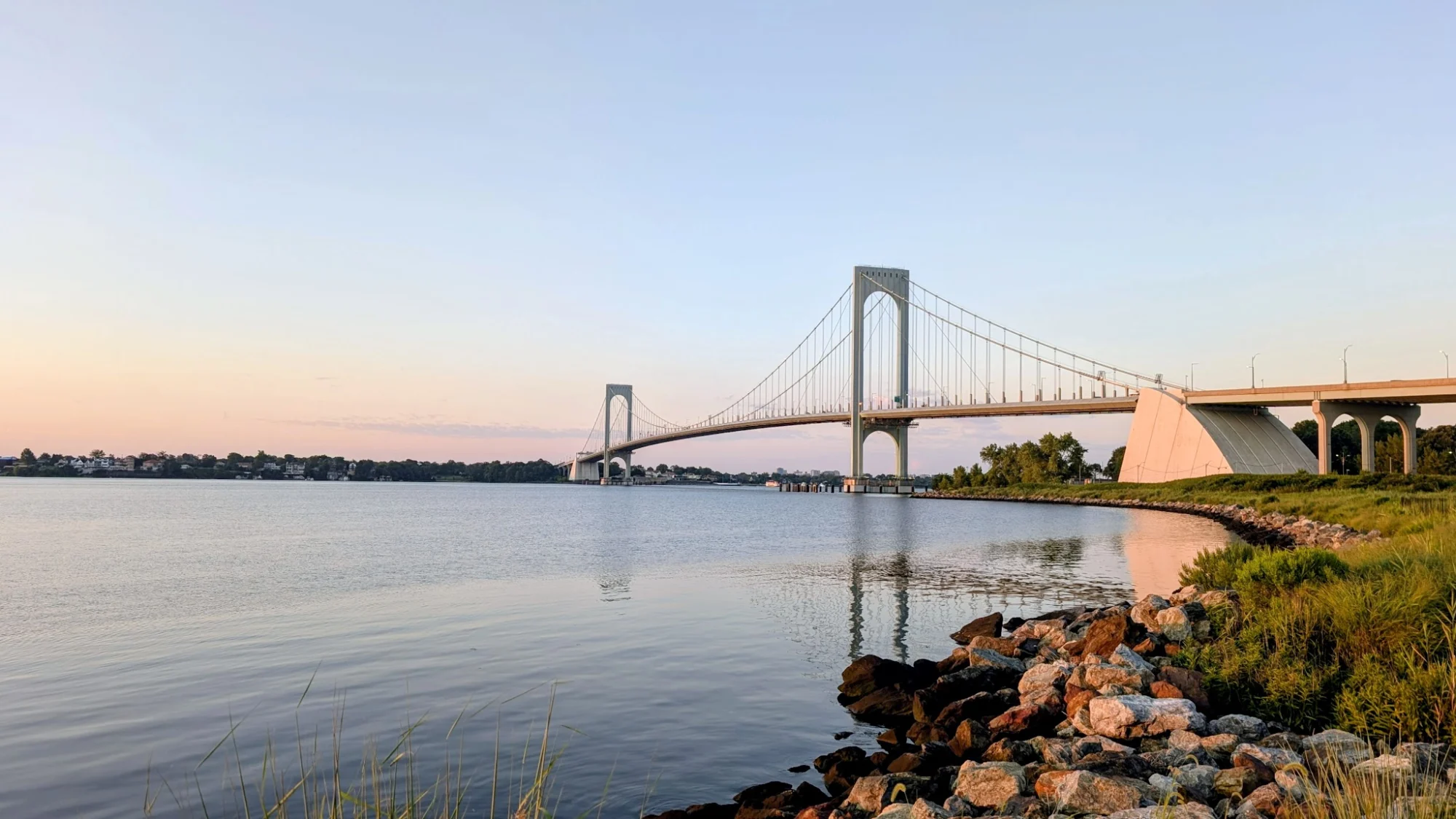 Wide view of a suspension bridge over a body of water with a rocky shoreline at Ferry Point Park, Throgs Neck.