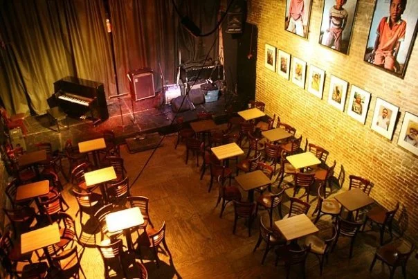 Nuyorican Poets Cafe featured image Interior of Nuyorican Poets Cafe with tables, chairs, and a stage in East Village, Bronx NYC.