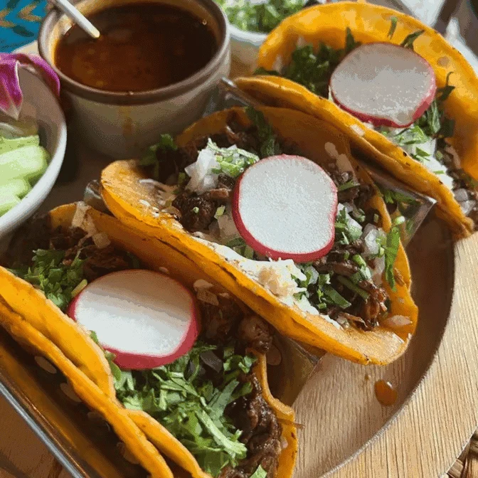 Three tacos filled with meat, lettuce, radish slices, and cheese on a wooden tray at Yolpaqui Taqueria, Port Richmond.