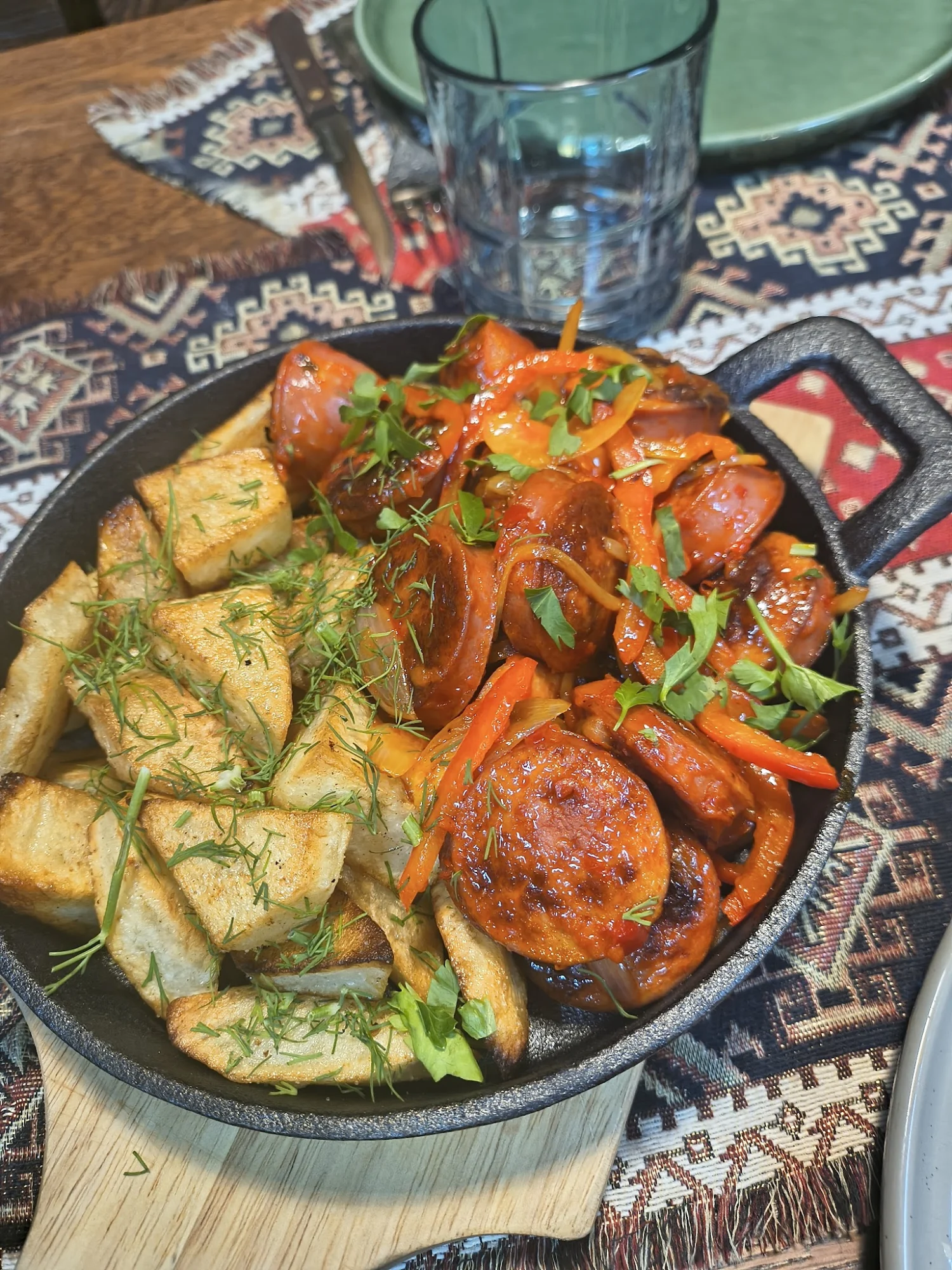 Sizzling skillet with sausages, fried potatoes, and herbs on a patterned tablecloth at Mr. Week, Arrochar.