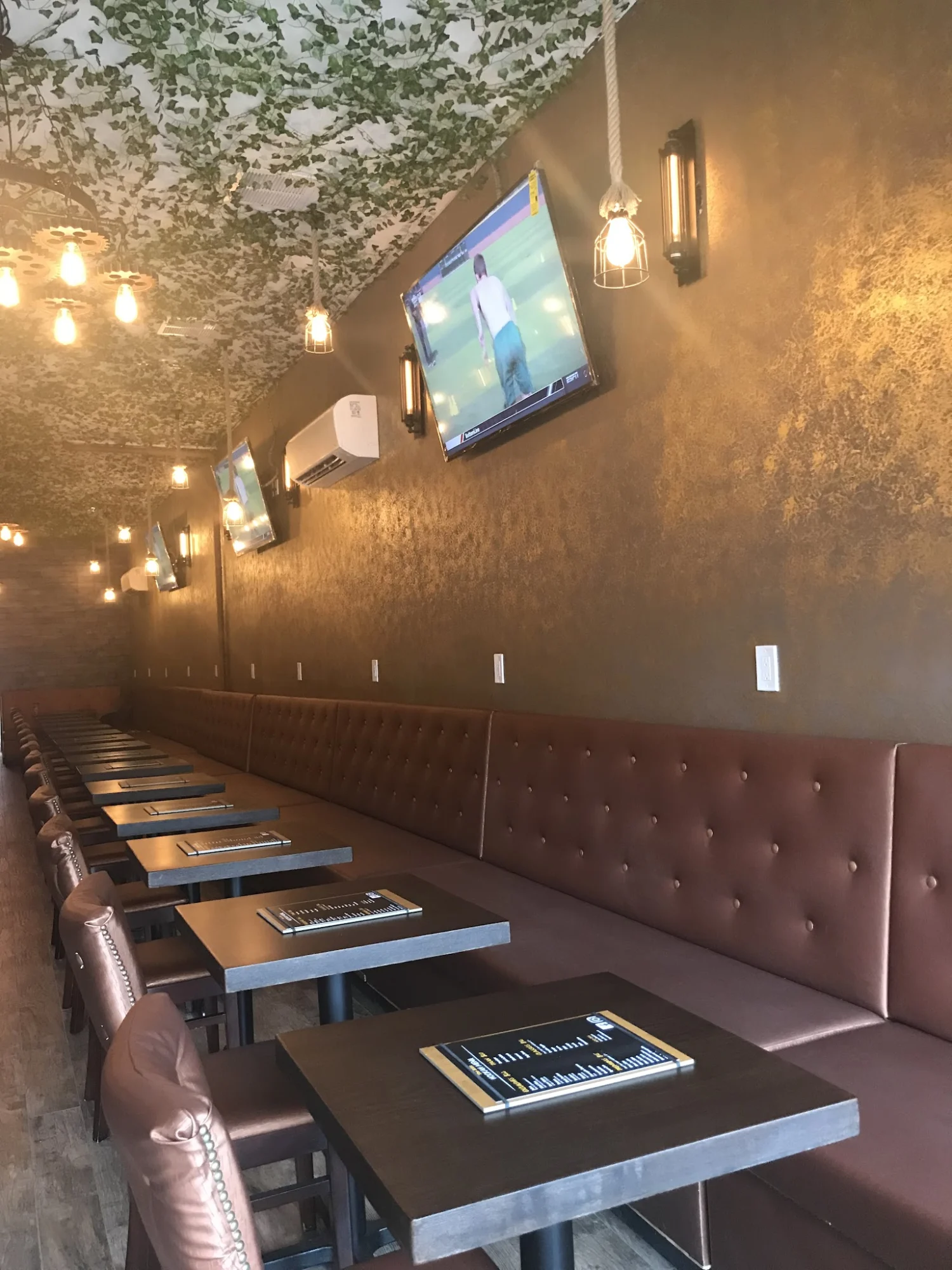 Interior of The Attic Hookah Cafe in Bay Ridge, Bronx NYC with row of tables and chairs, TV screens, hanging lights, and ivy ceiling.
