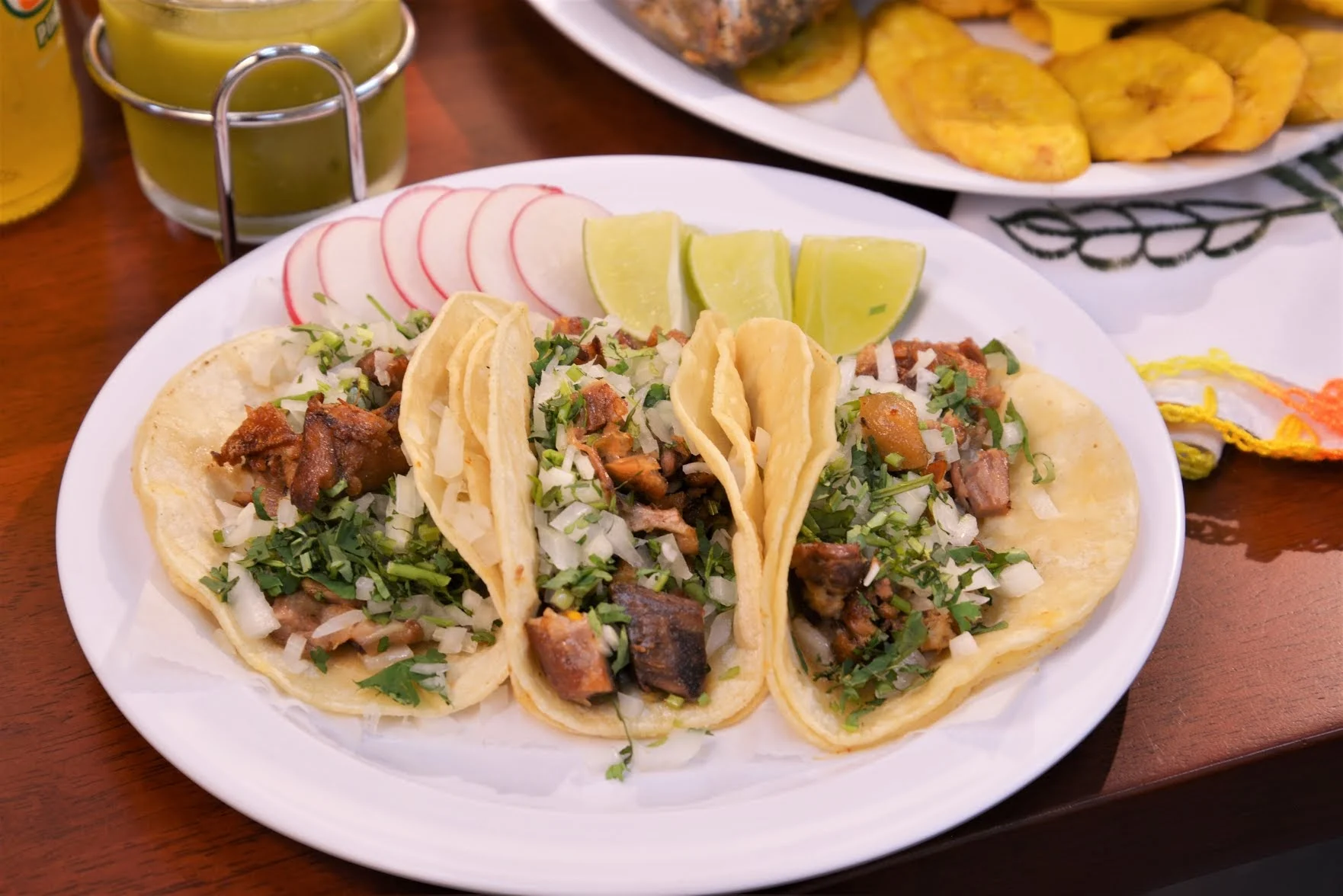 Three tacos with meat, cilantro, onions, lime wedges, and radish on a white plate at La Cocinita, Richmond Hill.