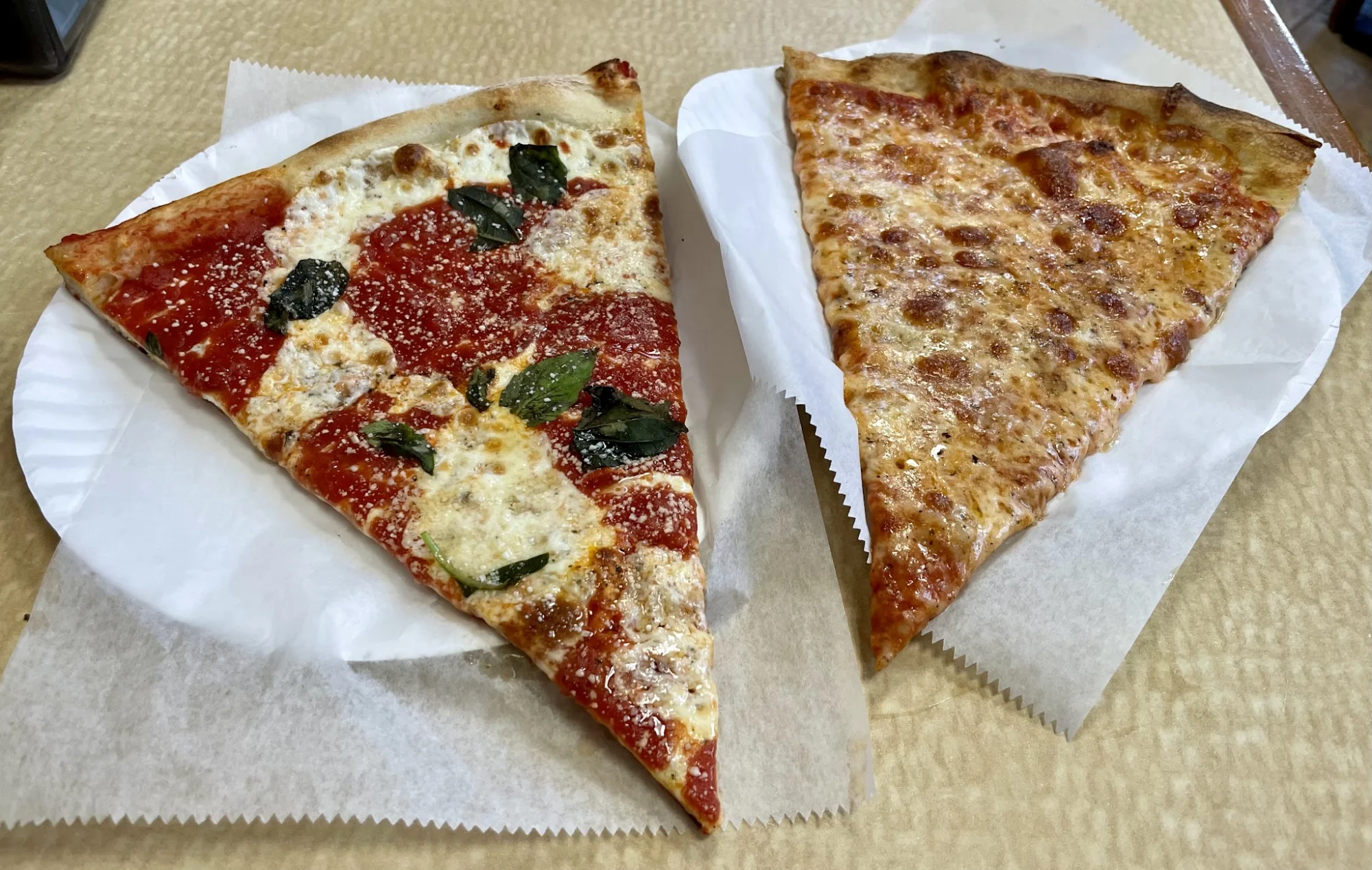 Two slices of pizza on paper plates, one with basil and cheese, on a wooden table at Louie & Ernie's Pizza, Pelham Bay.