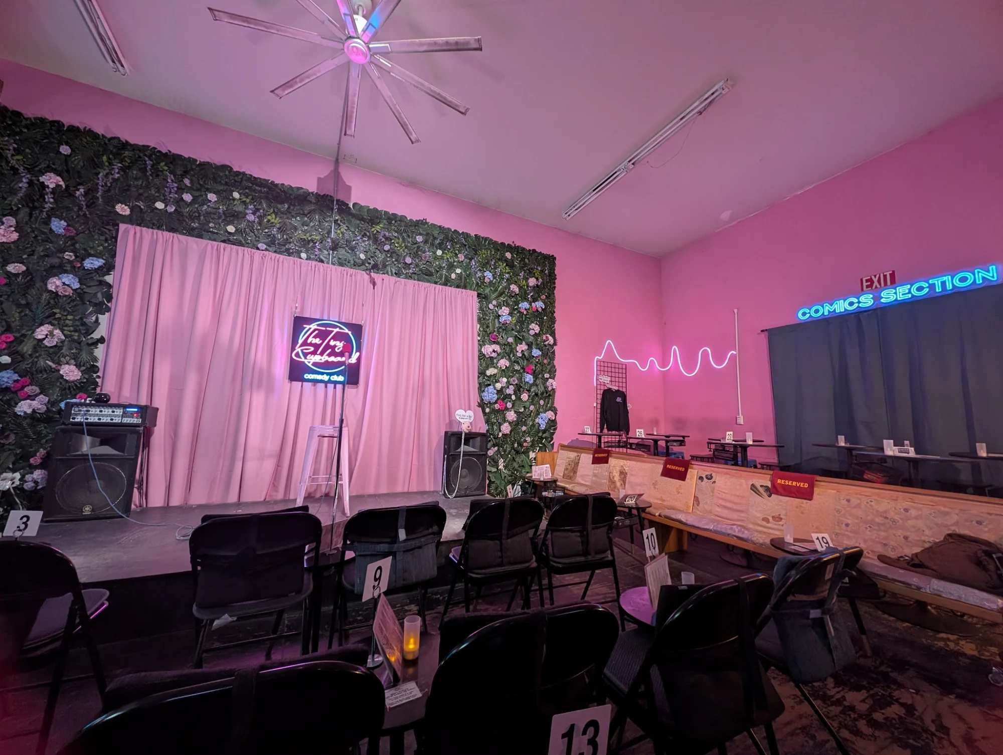 The Tiny Cupboard Comedy Club & Board Game Bar featured image Interior of The Tiny Cupboard Comedy Club with a stage, pink curtains, and 'Comics Section' neon in Bushwick.